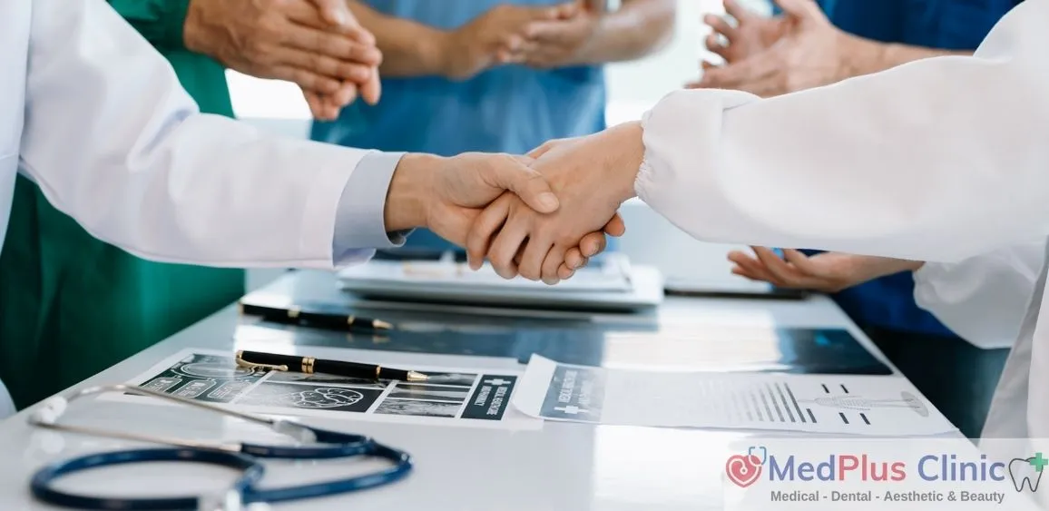 Two healthcare professionals at MedPlus Clinic shaking hands over a desk with medical documents and equipment, including stethoscopes and X-rays. Other medical staff are visible in the background, emphasizing collaboration and teamwork. The clinic's logo and services (Medical, Dental, Aesthetic & Beauty) are displayed in the bottom right corner.