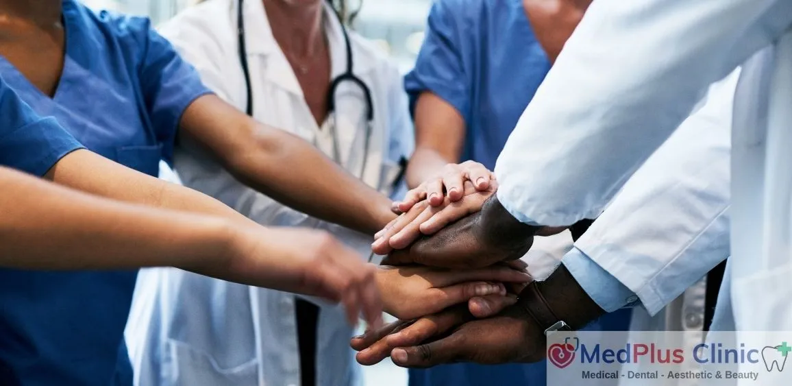 A diverse team of healthcare professionals at MedPlus Clinic joining hands in a show of unity and teamwork. The team includes individuals wearing scrubs and lab coats, some with stethoscopes, emphasizing collaboration and commitment to patient care. The clinic's logo and services (Medical, Dental, Aesthetic & Beauty) are visible in the bottom right corner."